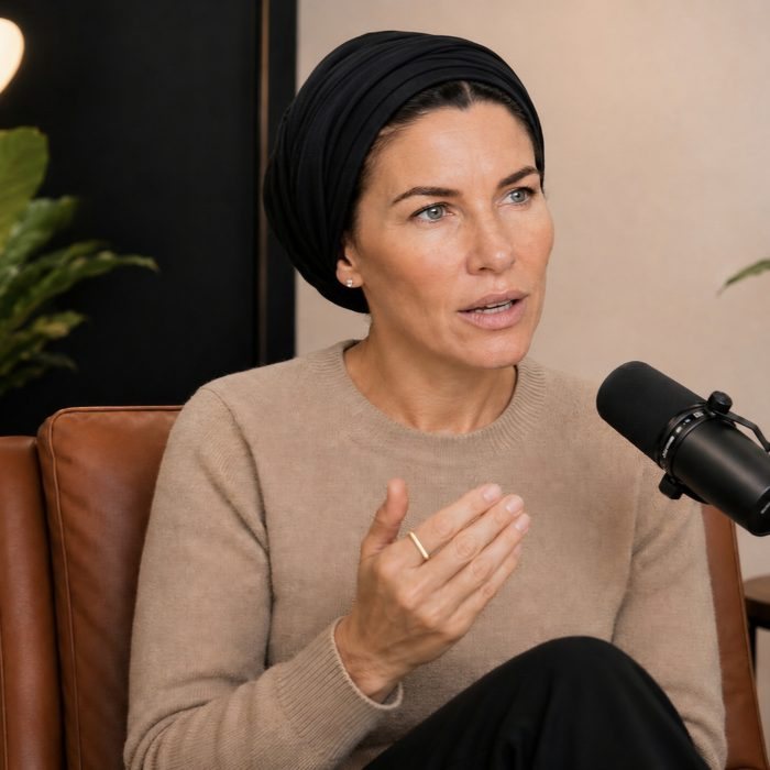 Rabin Stone mid conversation at a podcast microphone, wearing a black head wrap and a beige sweater, seated in a leather chair.